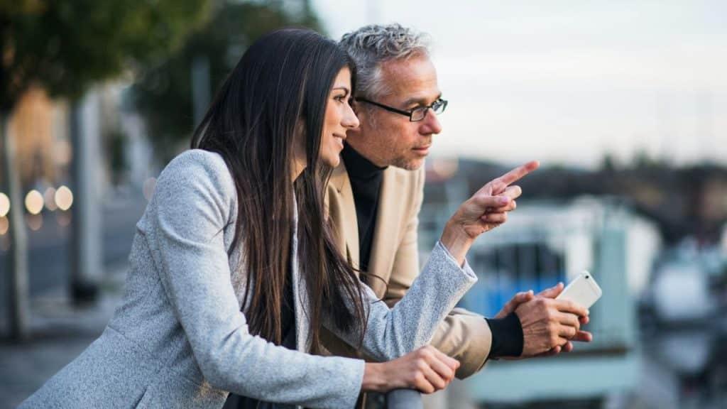 A man and woman lean against a railing outdoors, looking and pointing toward the distance.