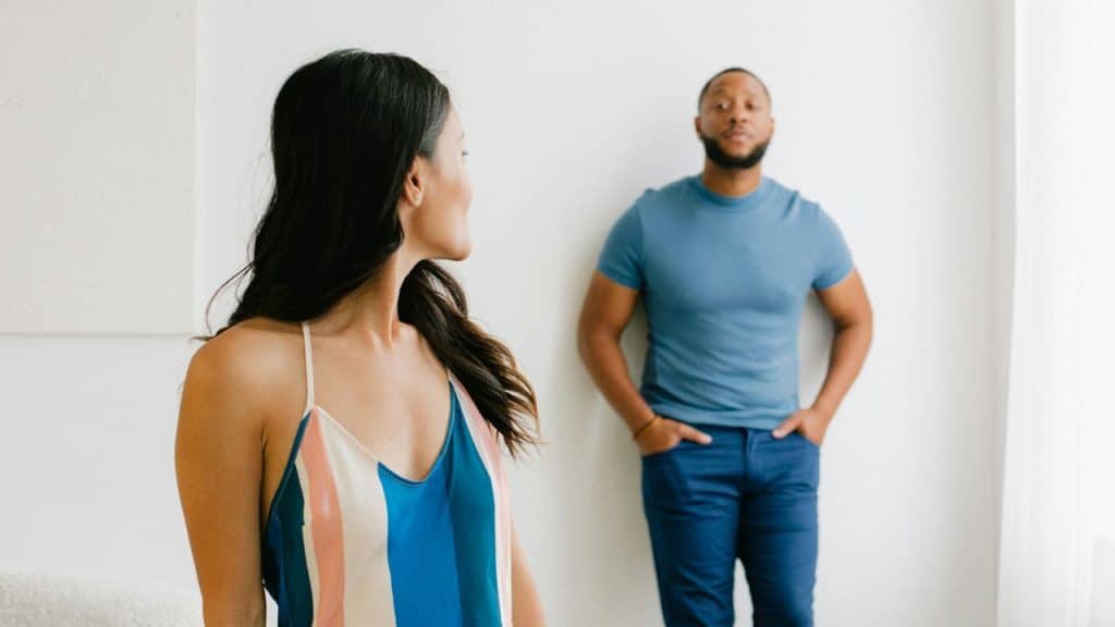 A woman in the foreground looks over her shoulder at a man leaning against a wall.