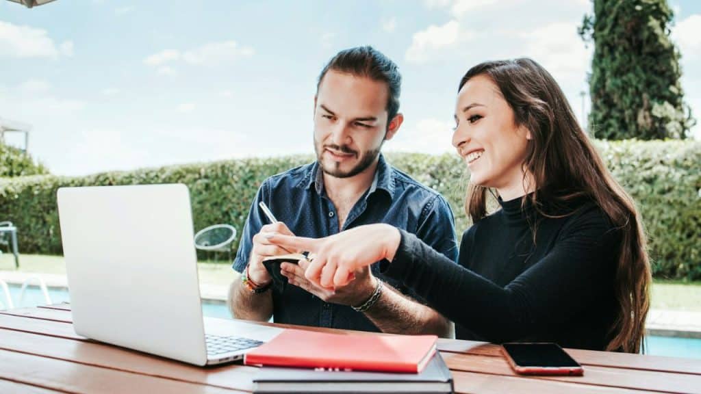 A man and woman sit at an outdoor table looking at a laptop computer together.