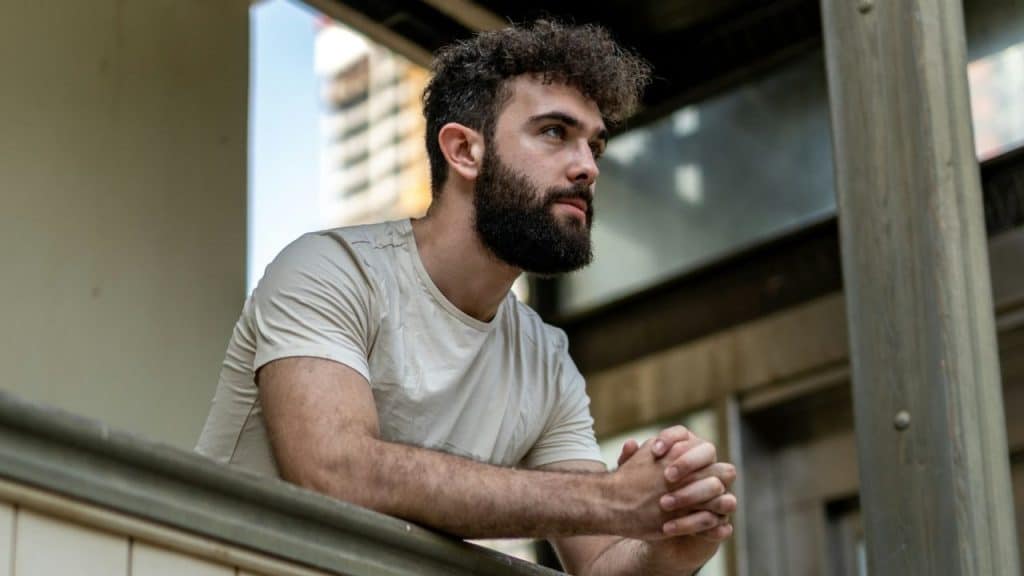A bearded man with curly hair leans on a railing, looking out with hands clasped.