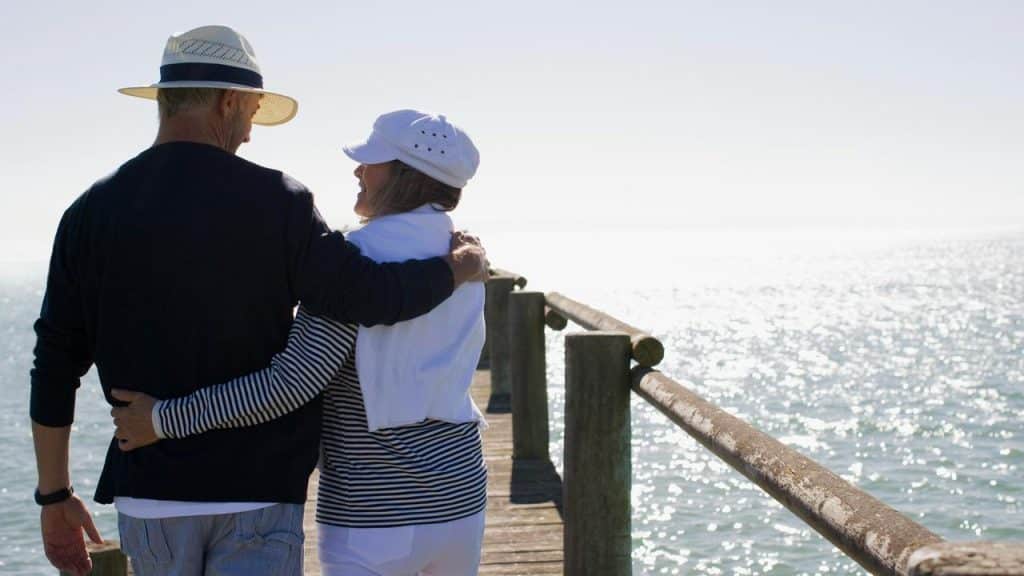 An elderly couple walking arm in arm on a sunny pier by the sea.
