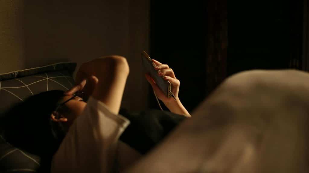 A person lying in bed at night holding a smartphone in dim light.