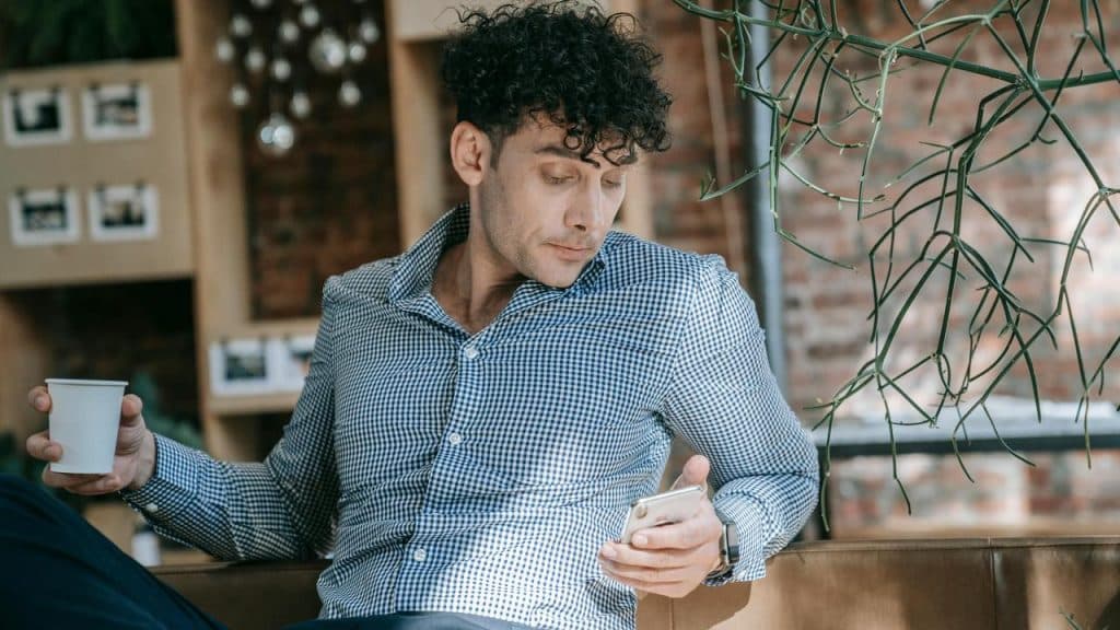 A man sitting indoors holding a cup and looking at his smartphone.