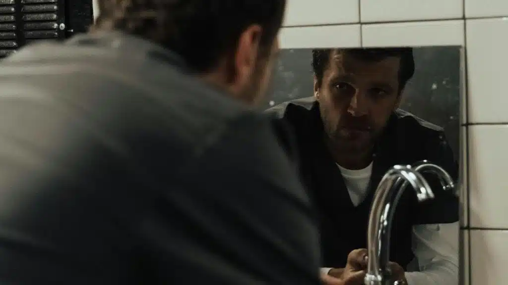 A man staring at his reflection in a dimly lit bathroom mirror.