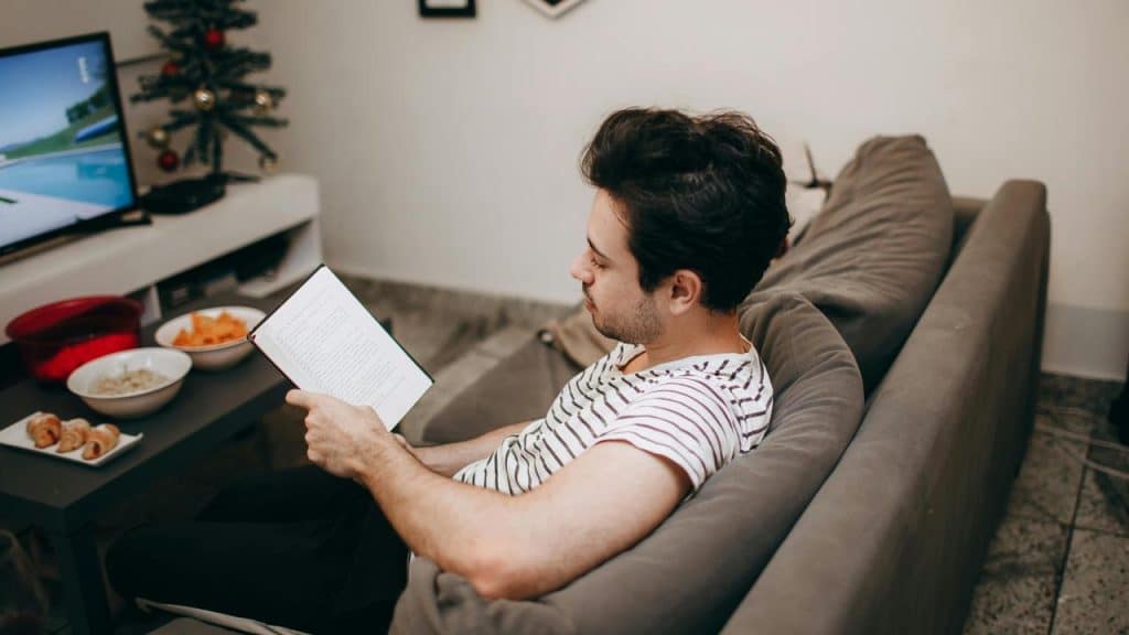 A man sitting on a couch reading a book near a table with snacks.