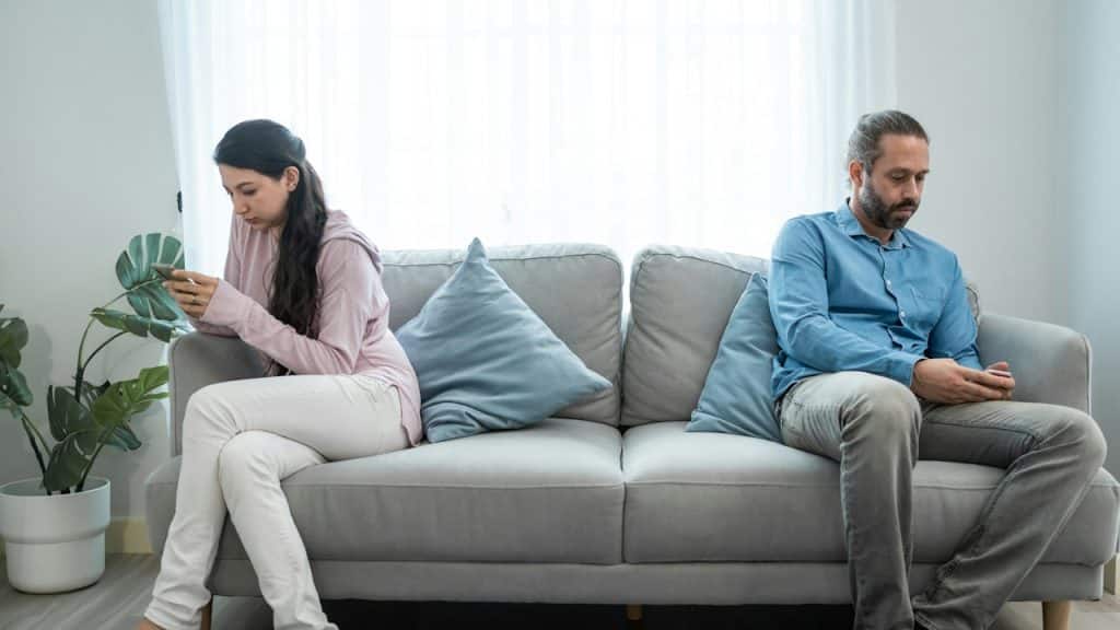 A couple sitting apart from each other on a couch and using their phones.