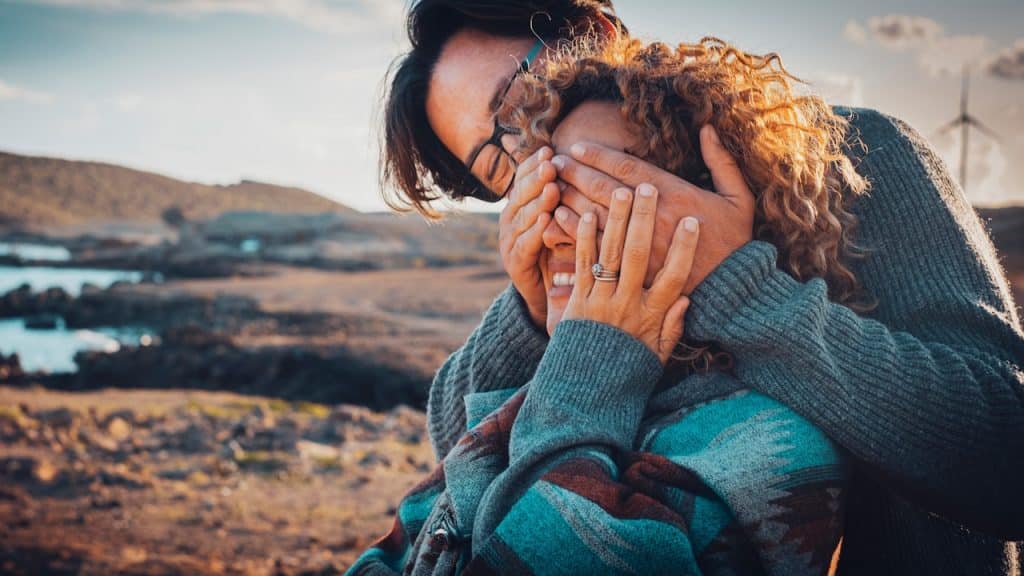 A man covering a woman’s eyes as she smiles while they stand outdoors.