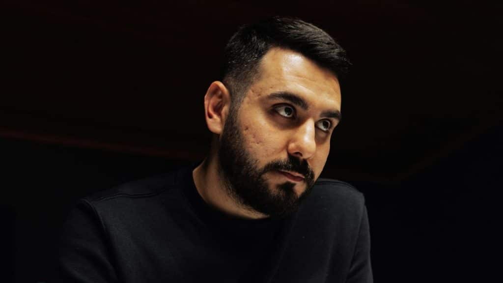 A bearded man in a black shirt looks toward the side against a dark background.
