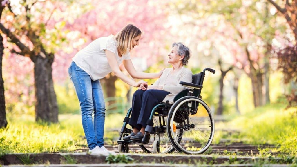 A woman stands in a park, smiling and holding the hands of an elderly woman in a wheelchair.