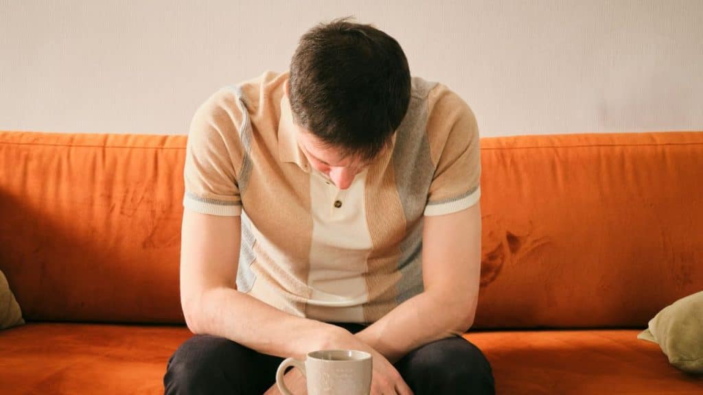 A man in a striped shirt sits on an orange sofa with his head bowed down.