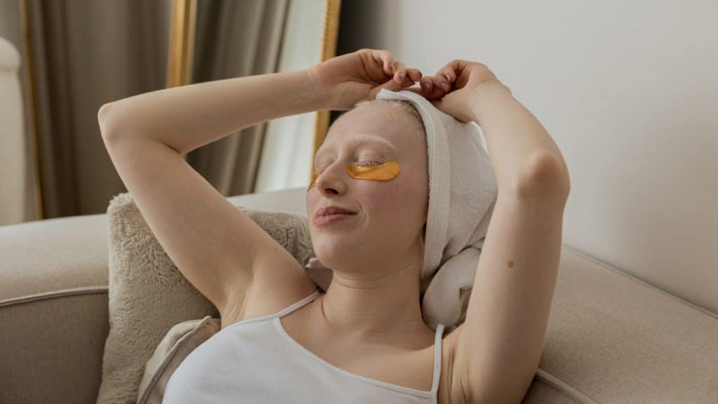 A woman with closed eyes wearing a towel on her head and skin cream under her eyes reclines on a couch and smiles.