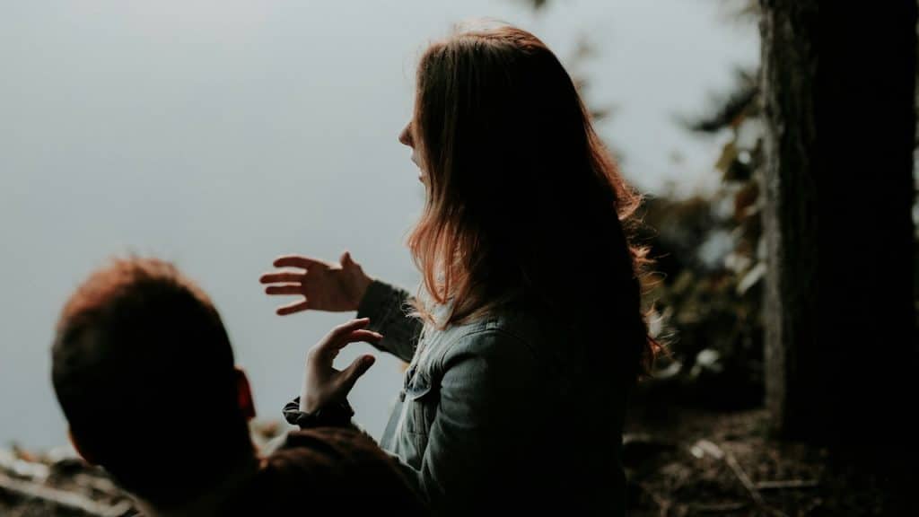 A woman gestures with her hands while speaking to a man in a hazy outdoor setting.
