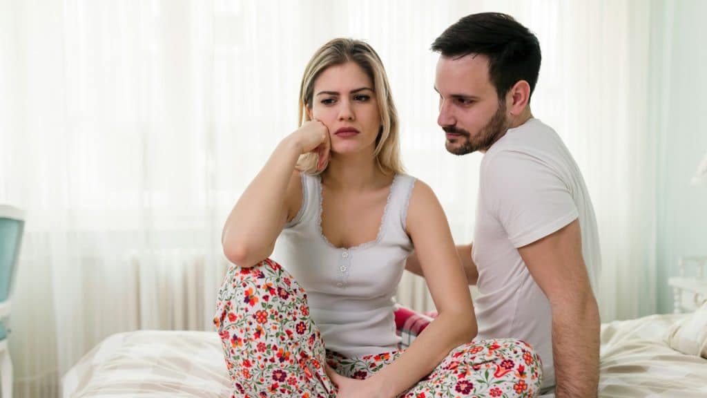 A blonde woman with a distressed expression is sitting cross-legged on a bed, while a man sits beside her looking at her with concern.