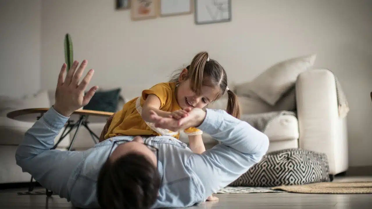 Father lying on the floor while his smiling young daughter plays on top of him.