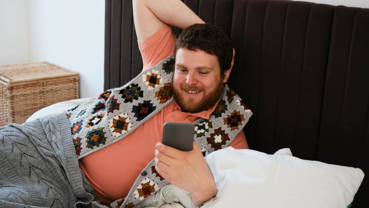 Man smiling while relaxing in bed, wrapped in a blanket and looking at a smartphone.