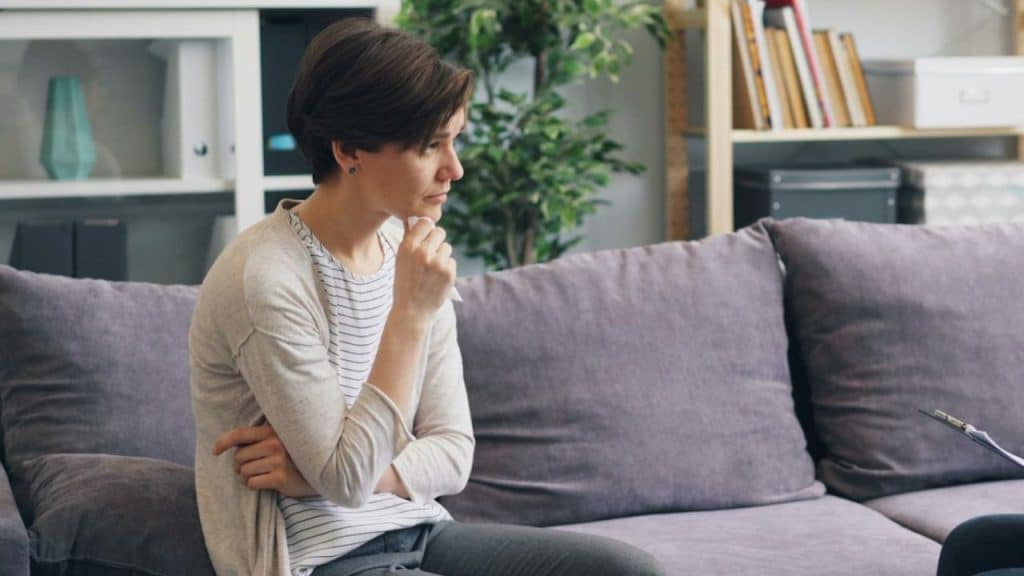 Woman with short hair sits on a gray couch, looking away thoughtfully, holding a tissue.