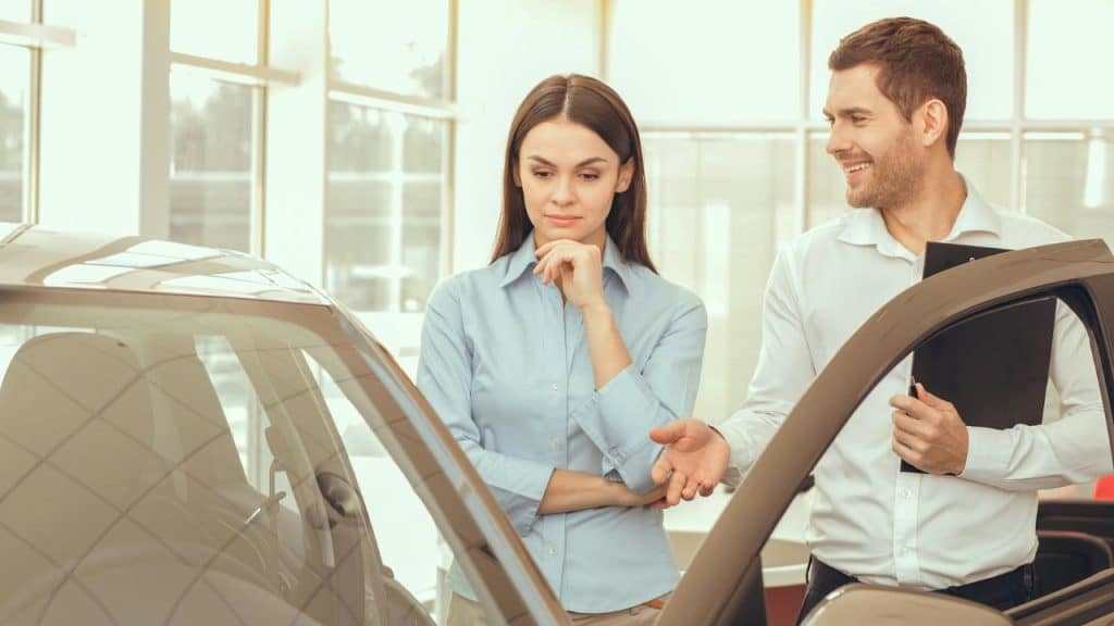 A woman examines a car while a man in a white shirt gestures at the open door.