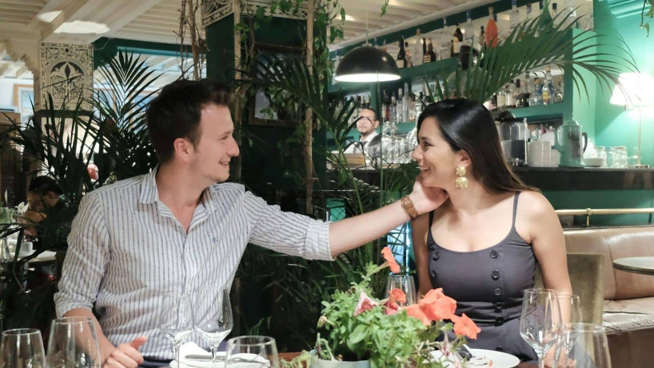 Couple smiling at each other during a dinner date in a restaurant with dark green and plant decor.