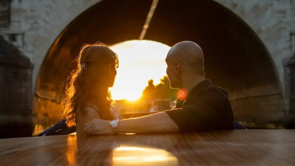 A man and a woman sitting in front of a bridge at sunset.