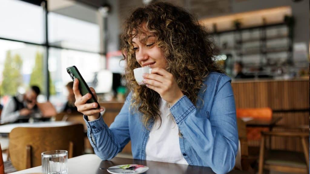 A happy young woman using a mobile phone and enjoying coffee at a cafe.