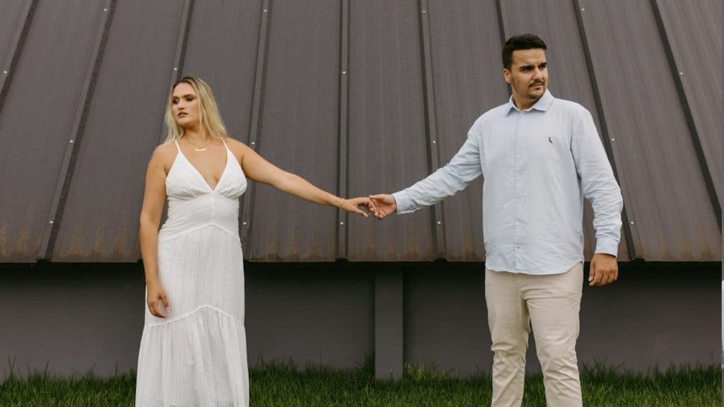 A couple holding hands yet standing apart and looking in different directions. 