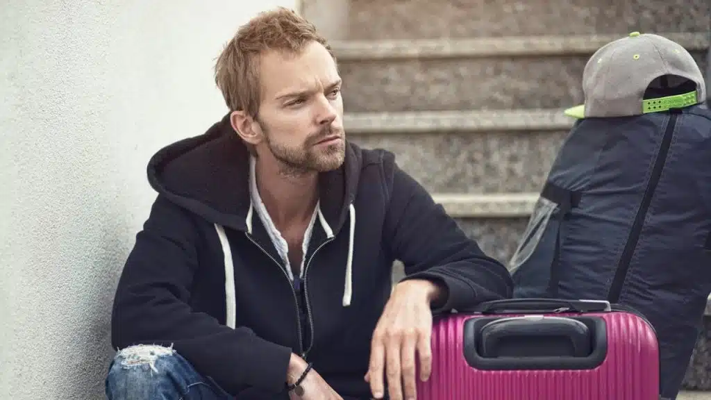 A lightly bearded man resting his arm on a suitcase and sitting on some stairs is looking to the side. 