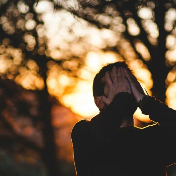 A silhouetted person covering their face at sunset among trees.
