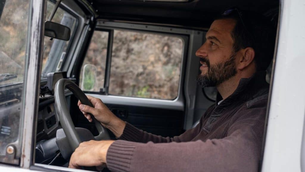 A bearded man driving a rugged vehicle and looking ahead through the windshield.