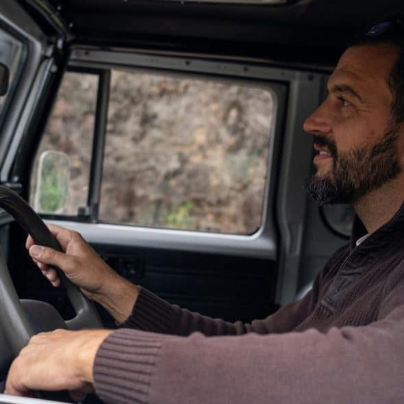 A bearded man driving a rugged vehicle and looking ahead through the windshield.
