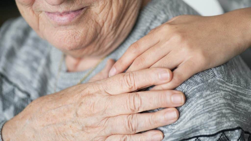 A close-up of an elderly person smiling while another hand rests gently on their shoulder.