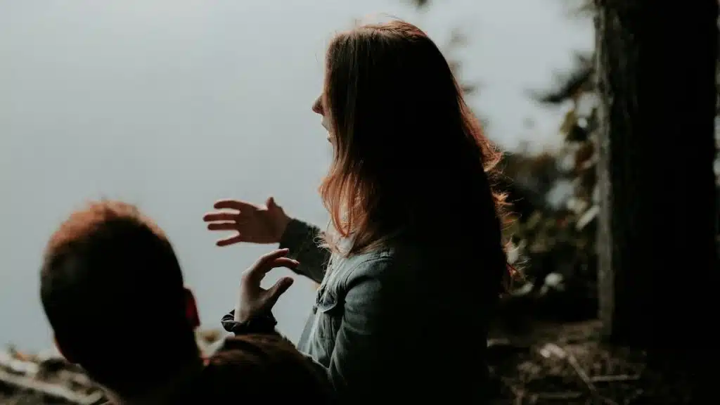 Woman gesturing with her hands while talking to a man outdoors by a foggy lake.