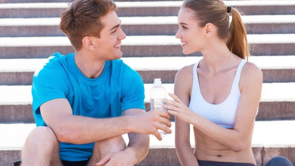 A couple smiling at each other while the man hands the woman a water bottle.