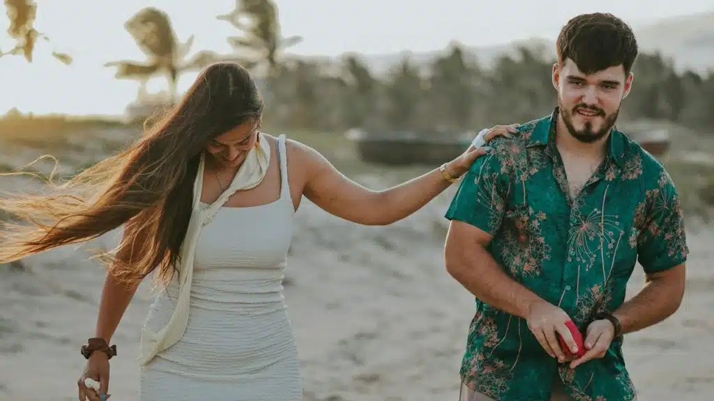A couple smiling and walking down a beach while the woman places her hand on the man’s shoulder.