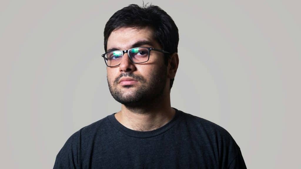 A solemn man with glasses and a beard stares at the camera.