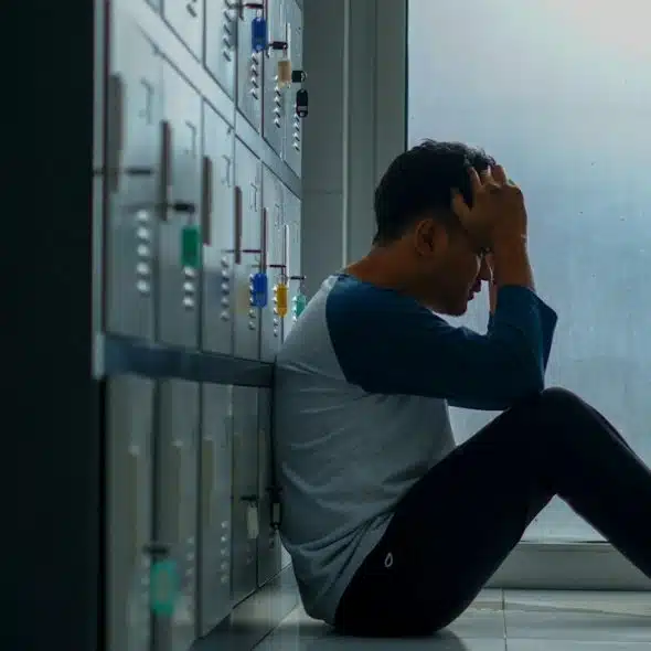 A man sitting on the floor while leaning on some lockers while holding his head in his hands.