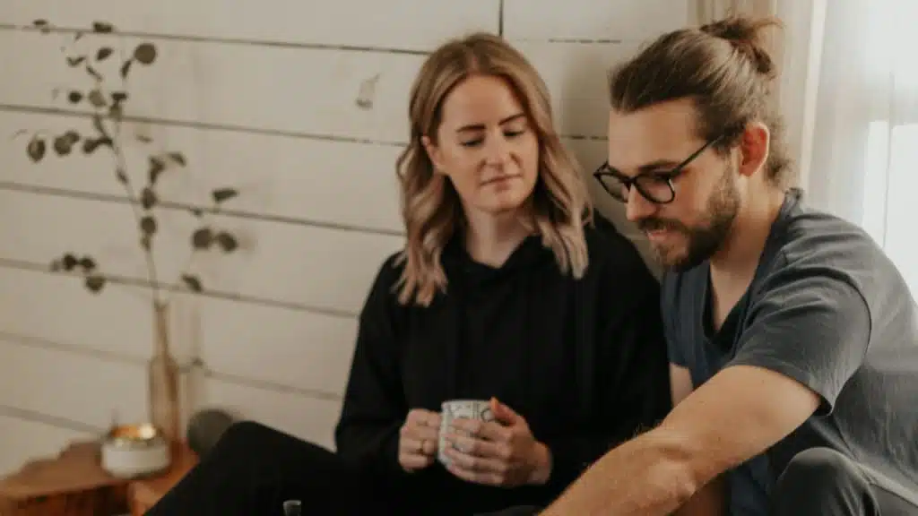A serious-looking couple sits on the floor, and the woman is holding a mug.