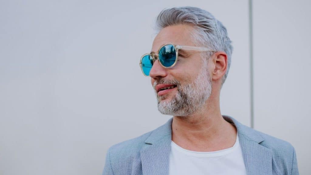A smiling, stylish older man with white hair and a beard looks to the side while wearing sunglasses.