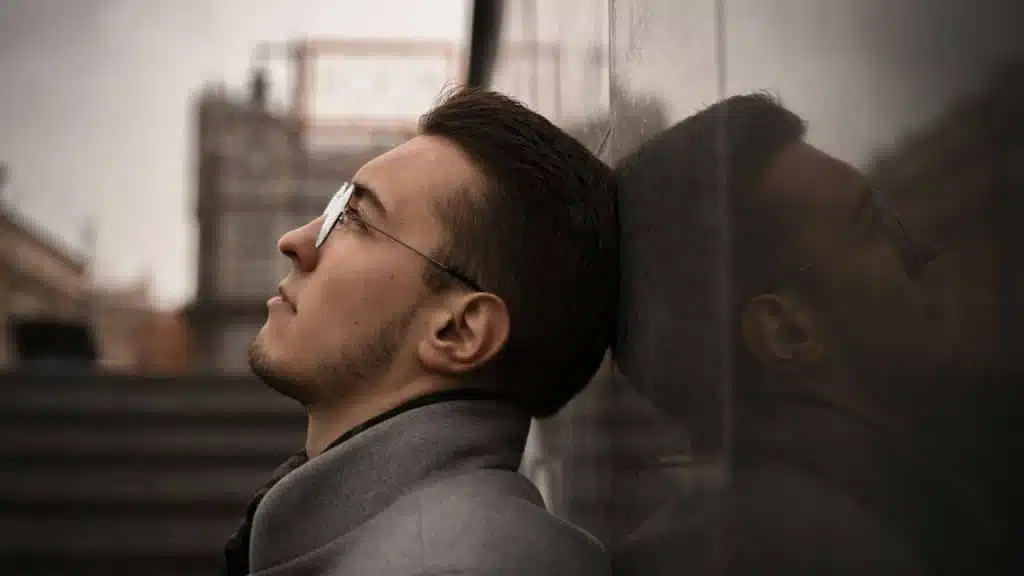A serious man with glasses and a beard is leaning his head against a stone wall, looking up.