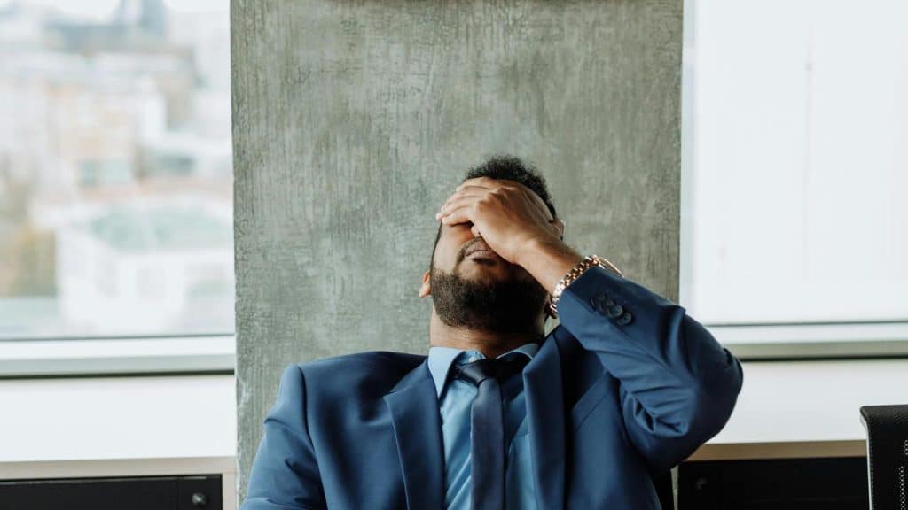 A person in a suit leans back in a chair with a hand covering their face in an office setting.