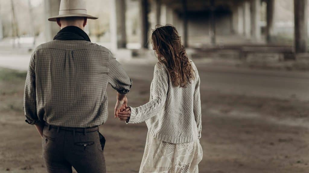 A man wearing a hat not holding a woman’s outstretched hand as they walk down a path outdoors.