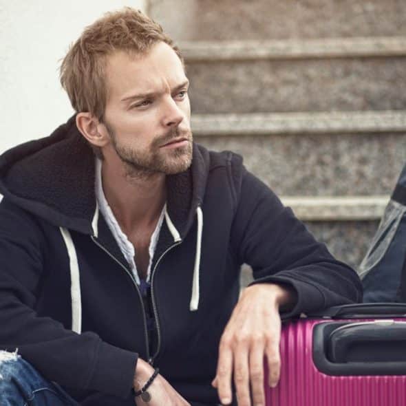 A lightly bearded man resting his arm on a suitcase and sitting on some stairs while appearing lost in thought.