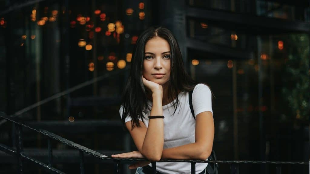 A woman holding her face with her hand and looking at the camera while leaning on a fence. 