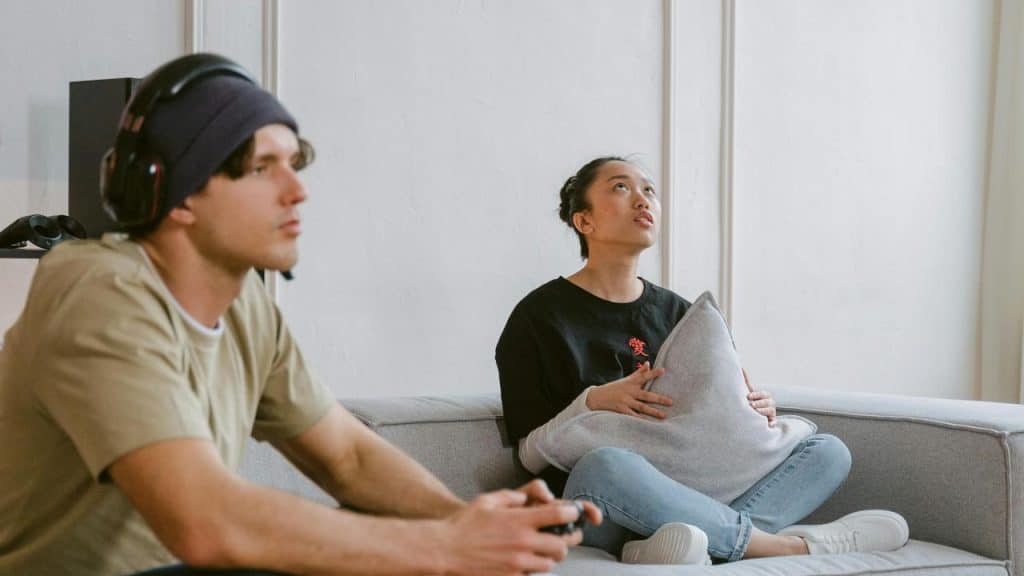 A person plays a video game while another sits on a couch holding a pillow.