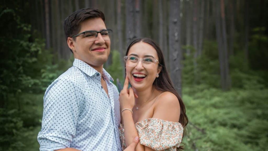 A couple wearing glasses standing close together in front of trees.
