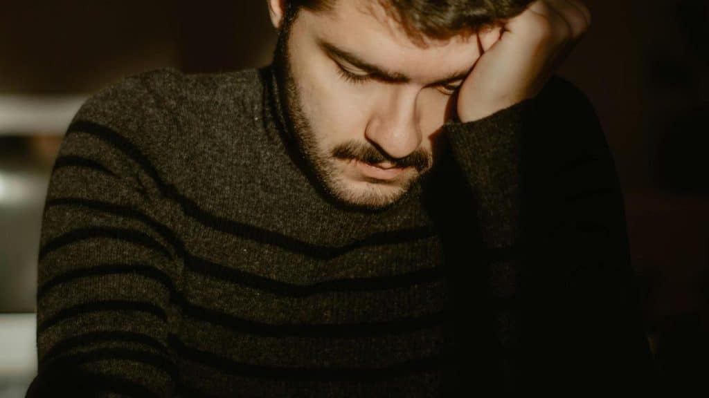 A person rests their head on one hand while looking down in soft light.