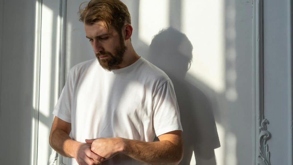 A person stands by a wall in sunlight, looking down with hands together.