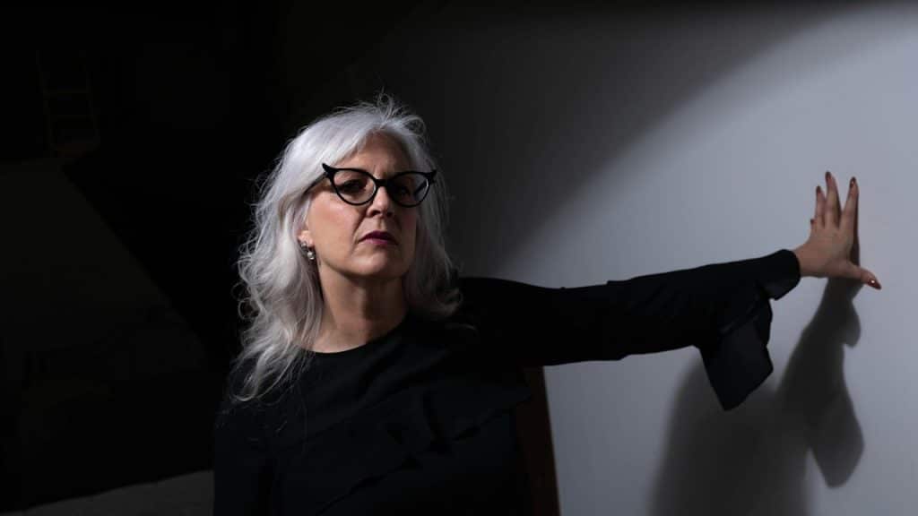 A mature white-haired woman wearing glasses and leaning against a wall.