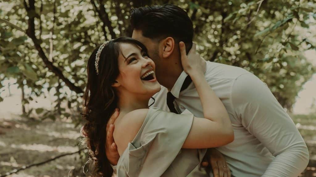 A man kissing laughing woman on the cheek while holding her close. 