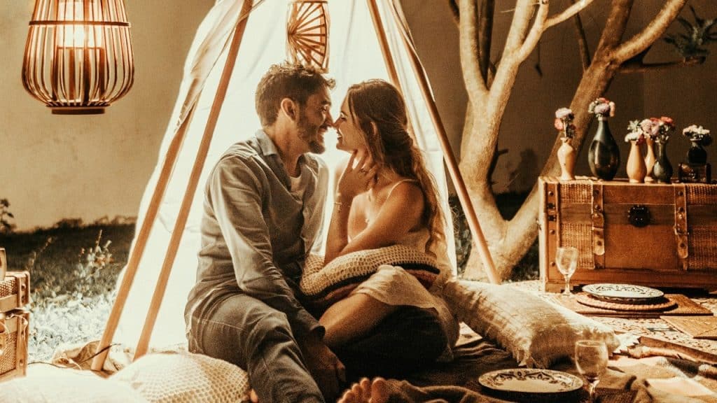 A smiling man and woman sitting under a makeshift tent in their living room and about to kiss.