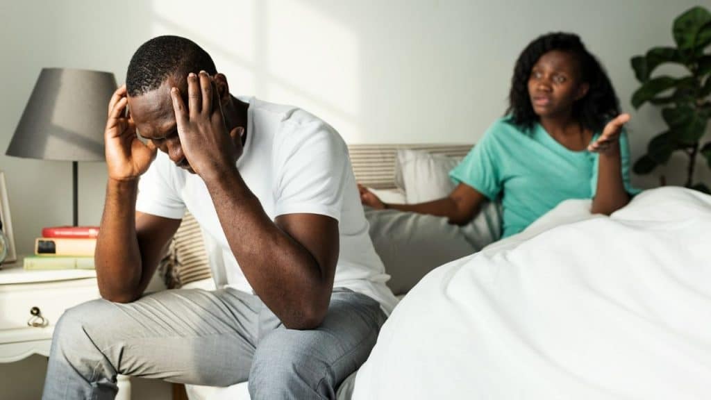 A Black man holds his head in distress as a Black woman argues with him in the background.